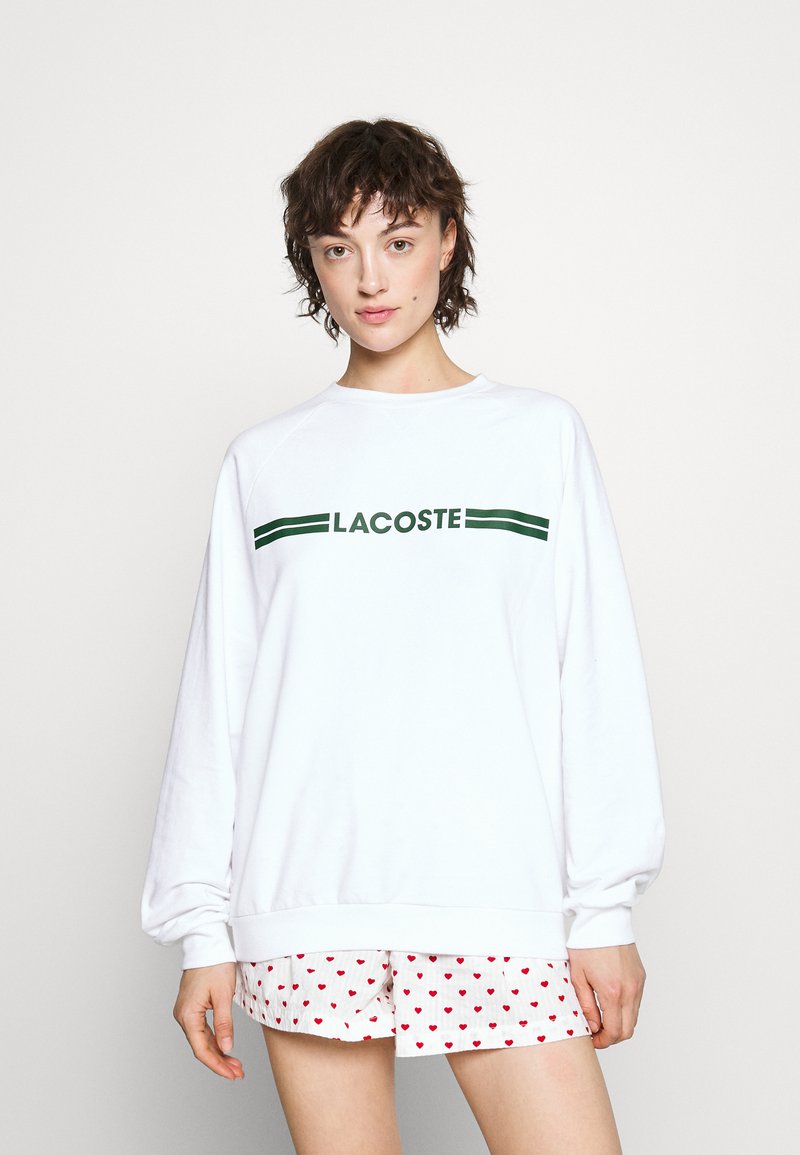 Witte sweatshirt met groene "LACOSTE" tekst, ronde hals, verlaagde schouders en geribbelde manchetten, gecombineerd met gestreepte rode hartshorts.