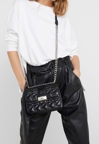 Borsa a tracolla nera trapuntata in faux leather, con borchie argentate e una chiusura rettangolare argentata. La tracolla a catena aggiunge un dettaglio di texture.