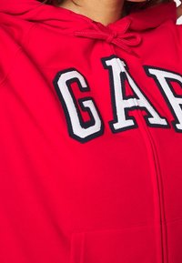 Sudadera roja hecha de una tela suave que presenta un gran logo bordado de "GAP" en blanco con contornos en azul marino. Equipado con una capucha y un bolsillo delantero.