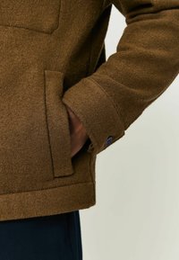 Hand in der Tasche einer braunen Wolljacke mit dunkelblauen geknöpften Ärmeln, über dunklen Hosen, vor einem schlichten hellen Hintergrund.