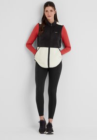 Black and cream fleece vest with a high collar, front zipper, and side pockets, paired with a red long-sleeve top and black leggings.