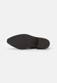 Chaussure en cuir marron avec un bout pointu, présentant un dessus lisse et une semelle en caoutchouc noir texturé avec une légère courbe au niveau du talon.