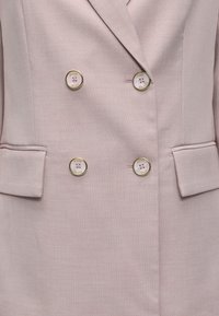 Blazer beige croisé à quatre boutons ronds arborant la marque "GUESS" et deux poches avant à rabat.
