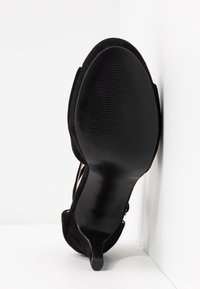 Czarne zamszowe buty na wysokim obcasie widziane od dołu, z gładką podeszwą z rowkami i szpiczastym noskiem.