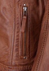 Braune Lederjacke mit Detail eines Reißverschlussfachs aus silbernem Metall und strukturiertem Oberflächenmaterial. Genähte Akzente vervollständigen das Design.