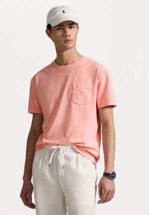 Helles Korallenfarbenes T-Shirt mit kurzen Ärmeln und Tasche, kombiniert mit beigen Jogginghosen mit Kordelzug. Eine weiße Mütze und eine braune Uhr runden das Outfit ab.