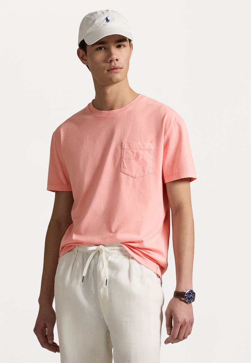 Light coral short-sleeve t-shirt with a pocket, paired with beige drawstring trousers. A white cap and brown watch complete the outfit.