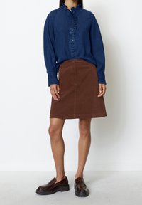 Denim shirt met een gerimpelde kraag, een bruine A-lijn rok en donkere instappers, wat een casual en gelaagd outfit benadrukt. Effen kleurenschema met textuur.