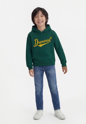 Băiat zâmbitor purtând hanorac verde cu text galben „Dsquared²”, blugi albaștri și adidași albi, stând pe fundal alb.
