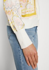 Kardigan w kwiatowy wzór w stonowanym różu, żółci i szarości. Żeberkowe mankiety i biały korpus. Posiada strukturalny splot i zapięcie na guziki.