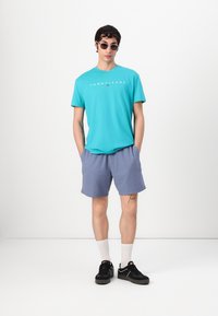 Tyrkysové bavlnené tričko s logom "TOMMYJEANS", kombinované so svetlomodrými šortkami, čiernymi teniskami a slnečnými okuliarmi, umiestnené na jednofarebnom pozadí.