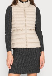 Beige Steppweste mit hohem Kragen, quiltierter Textur und goldenen Reißverschlussakzenten. Getragen über einem dunklen gerippten Pulloverkleid.