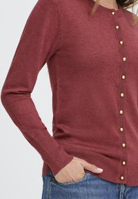Rote Strickjacke mit langen Ärmeln, mit goldenen Knöpfen auf der Vorderseite, gerippten Bündchen und Saum, getragen über blauen Jeans.