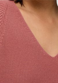 Gestrickter V-Ausschnitt-Pullover in sanftem Pink, mit strukturiertem Muster und lockerer Passform, der den Ausschnitt und den Schulterbereich betont.
