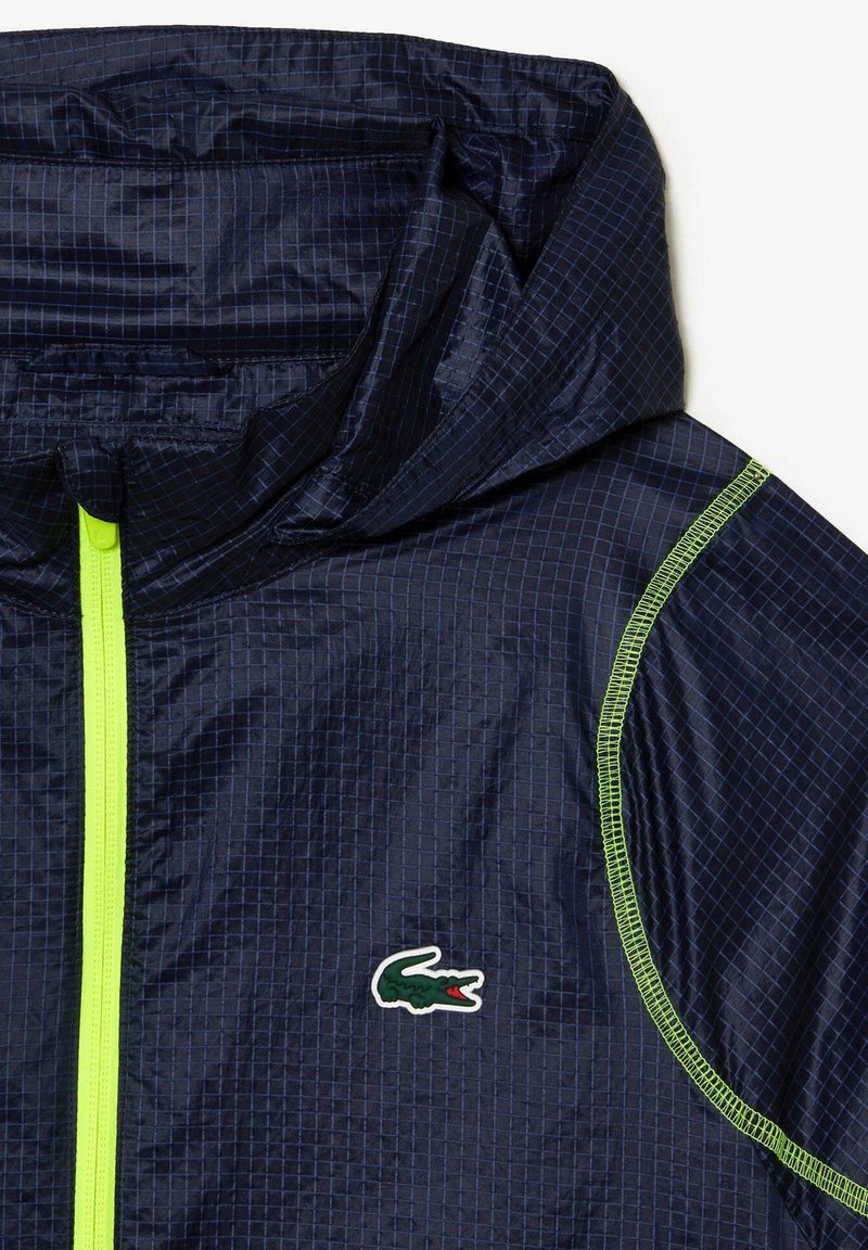 Marineblauer Windbreaker mit Karomuster, neongrünem Reißverschluss und Akzenten, strukturiertem Stoff und kleinem grünen Krokodil-Logo.