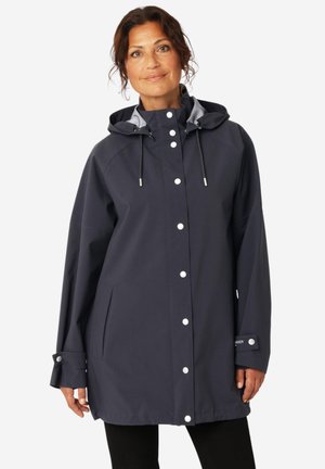 Woman wearing a navy blue hooded raincoat with snap buttons, front pockets, and adjustable cuffs, standing against a white background.