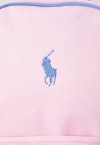 Mochila rosa con acabado texturizado, con un logo de jugador de polo bordado en azul. El diseño incluye acentos azules a lo largo de los bordes.