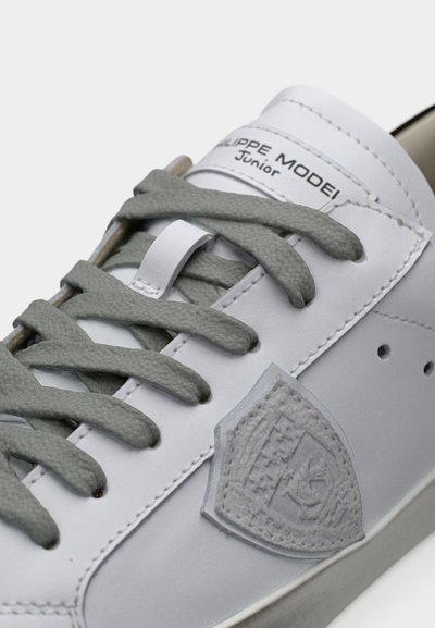 Baskets blanches avec lacets gris, un logo bouclier texturé gris sur le côté, et le texte "PHILIPPE MODEL Junior" sur la languette.
