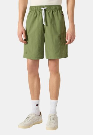 Uomo che indossa pantaloncini cargo verde oliva con coulisse bianca, calzini bianchi Champion e sneaker beige, in piedi su uno sfondo semplice.