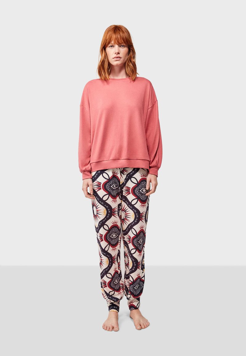 Sudadera rosa con hombros caídos y puños acanalados, combinada con pantalones crema que tienen un patrón geométrico en azul y rojo.