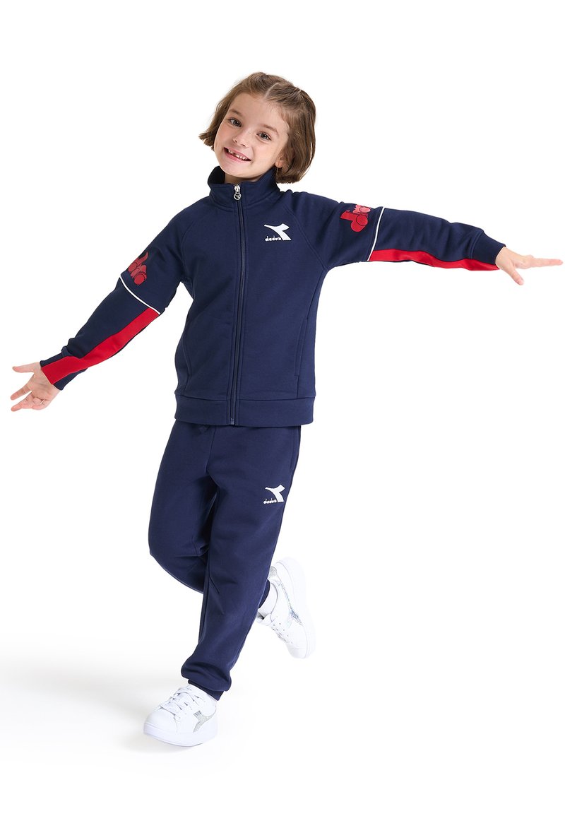 Tuta blu navy con giacca con zip e dettagli rossi, caratterizzata da un colore uniforme e un logo. Le sneakers bianche completano l'outfit.