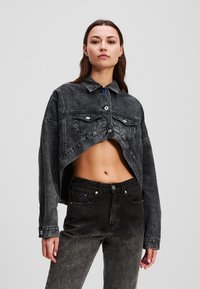 Veste en denim noir cropped avec un col et des boutons à l'avant, dotée de deux poches poitrine. Associée à un jean en denim noir taille haute.