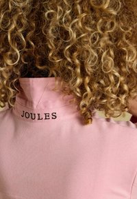 Heled roosa polo särk, millel on struktureeritud krae ja tikitud 'Joules' logo kaeluses. Kangas näib struktuurne ja sile.