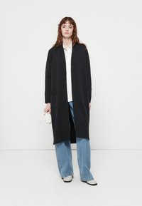 Jeune femme aux cheveux roux portant un long cardigan noir, une chemise blanche, un large jean bleu et des chaussures blanches, tenant un petit sac à main blanc devant un fond uni.
