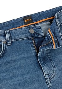 Modré džínové kalhoty s kovovým zipem, měděně tónovaným šitím a černou nášivkou s nápisem "BOSS" v oranžové barvě.