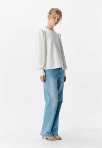 Weißer Pullover mit puffähnlichen Ärmeln, kombiniert mit hellblauen gerade geschnittenen Jeans; das Modell trägt beige Sandalen, vor einem schlichten Hintergrund.