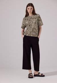 Oversized tričko s leopardím vzorem v béžové a černé barvě, spárované s černými širokými kalhotami. Modelka má na sobě ploché černé sandály se sponami.