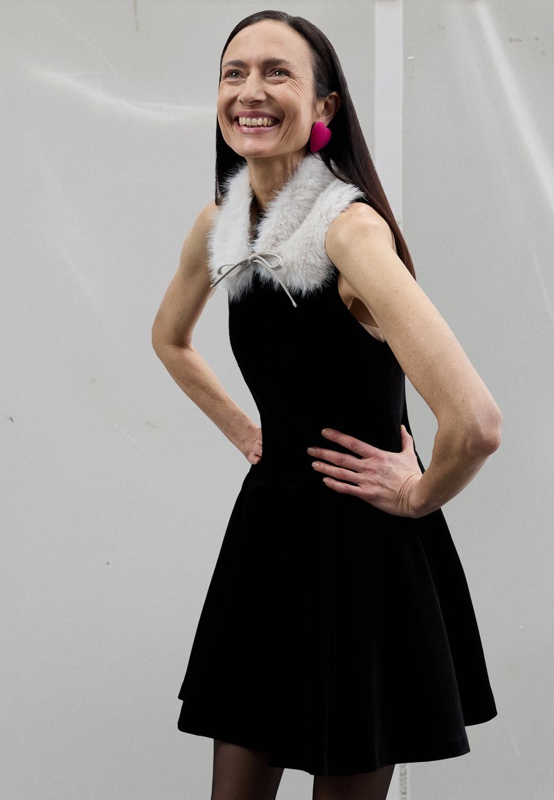 Zwarte mouwloze jurk met uitlopende rok, voorzien van een faux fur kraag en een strikdetail, gecombineerd met grote roze oorbellen in hartvorm.
