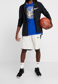 Černá lehká bunda s bílými akcenty, modré tričko s grafikou, krémové síťované šortky s černým lemováním a černé tenisky se zelenými akcenty. Drží basketbalový míč.