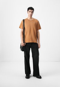 T-shirt en coton marron, coupe décontractée, associé à un pantalon en denim noir à jambes larges. Un sac à bandoulière noir et des chaussures noires épaisses viennent compléter le look.