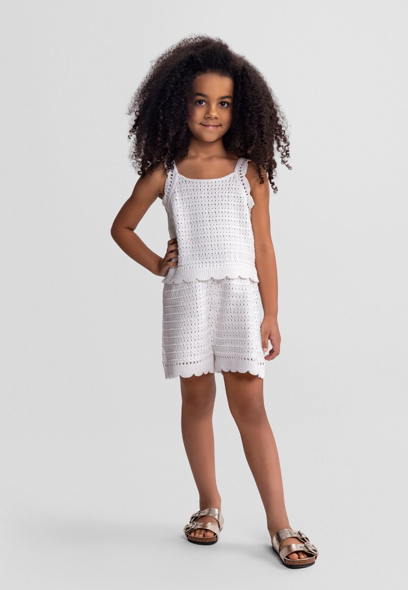 Jeune fille aux cheveux bouclés portant un débardeur blanc en crochet et un short, debout avec une main sur la hanche et des sandales métalliques.