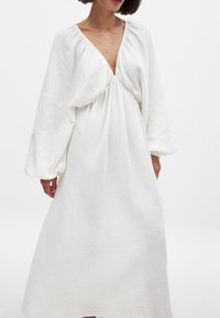 Weißes, langärmliges Kleid mit tiefem V-Ausschnitt und gerafftem Taillenbereich. Hergestellt aus strukturiertem, leichtem Stoff mit fließender Silhouette.