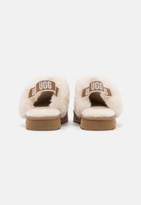 Pantoufles en beige flou avec une partie supérieure en fourrure douce, design à dos ouvert et une sangle avec le logo UGG en brun. Semelle plate avec un motif texturé.