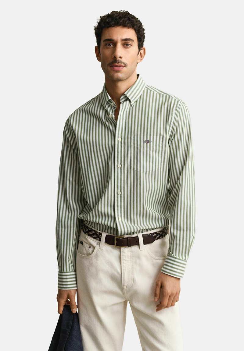 Uomo che indossa una camicia a righe verticali verdi e bianche infilata in pantaloni avorio con cintura marrone, che tiene un tessuto scuro nella mano sinistra.