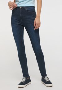 Jeans ajustados de denim azul oscuro con una textura suave, cinco bolsillos, cierre de botón y costuras sutiles a lo largo de las piernas. Combinados con zapatillas.