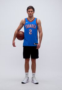 Joueur de basketball masculin portant un maillot bleu des Thunder, tenant un ballon de basketball sous le bras, debout sur un fond blanc uni.