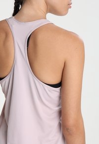 Top deportivo de color rosa claro con un diseño de espalda nadadora y textura transpirable. Sujetador deportivo negro visible debajo. Tejido suave.