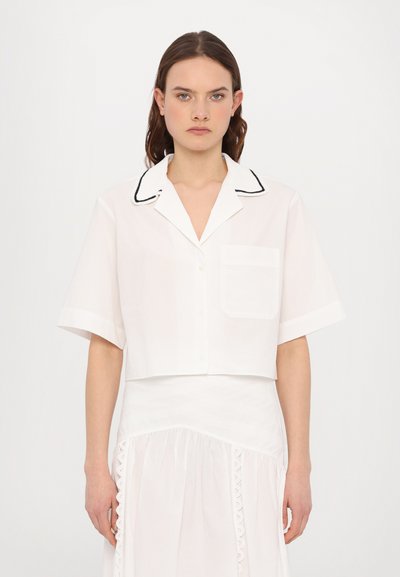 Hvid, kortærmet button-up skjorte med cropped længde, der har en sort kantet krave og en brystlomme. Stoffet ser let og glat ud.