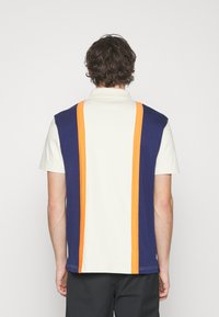 Polo à blocs de couleur bleu marine et crème, avec des rayures oranges dans le dos. Manches courtes et col classique. Matériau en mélange de coton.