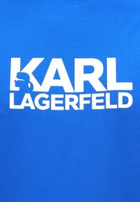 Zils kokvilnas T-krekls ar lielu baltu uzrakstu "KARL LAGERFELD" un silueta grafiku profilā burta "K" iekšpusē.