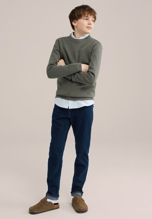 Olivengroen geribbelde trui, lichtblauw shirt eronder, donkerblauwe spijkerbroek en lichtbruine instappers. Casual outfit met gelaagde texturen.