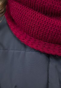 Gruba, dziergana bordowa szalik o teksturowanym wzorze, nałożona na gładką, szaro-puchową kurtkę. Udekorowana warkoczowymi brzegami.