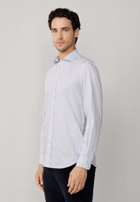 Hombre con cabello oscuro y rizado, vistiendo una camisa de cuadros azul claro de manga larga y pantalones oscuros, de pie frente a un fondo claro y liso.