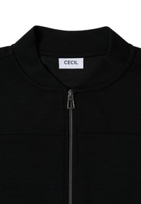 Veste zippée noire en tissu lisse avec un col montant et une fermeture éclair métallisée argentée, portant une étiquette avec la mention "CECIL."