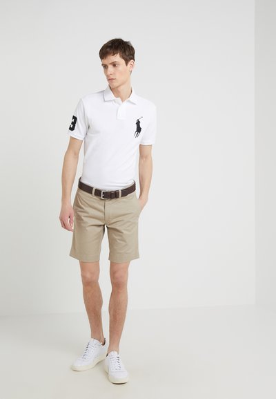 Biała koszulka polo z czarnym logo i numerem 3 na rękawie, w połączeniu z khaki szortami i białymi trampkami. Prosty, codzienny design.