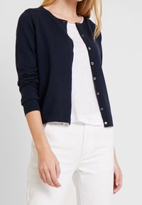 Marineblauer Cardigan aus weichem Material mit langen Ärmeln, rundem Ausschnitt und Knopfverschluss; kombiniert mit einem weißen Oberteil und hellen Hosen.